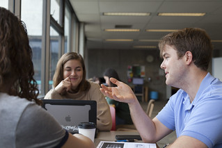 Students talking to each other at a cafe table. One has his hand out gesturing while another leans on her hand and look over a laptop, listening