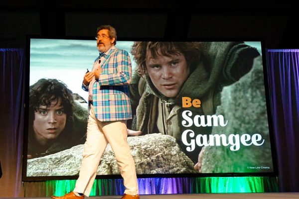 Presenter on stage in front of a slide showing an image from the movie LORD OF THE RINGS with the text BE SAM GAMGEE